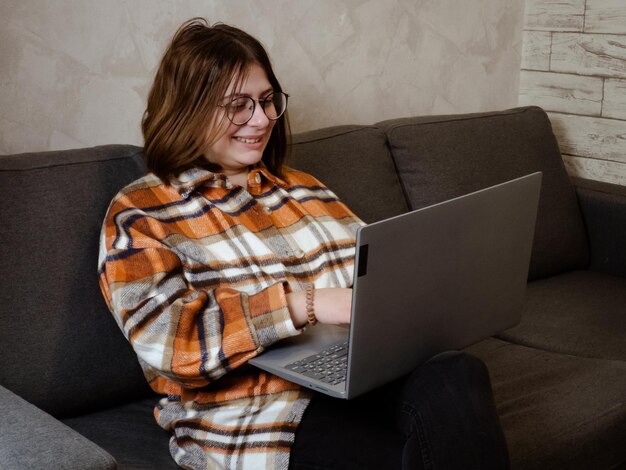 Jovem de óculos trabalhando em um laptop sentado em casa na cama.