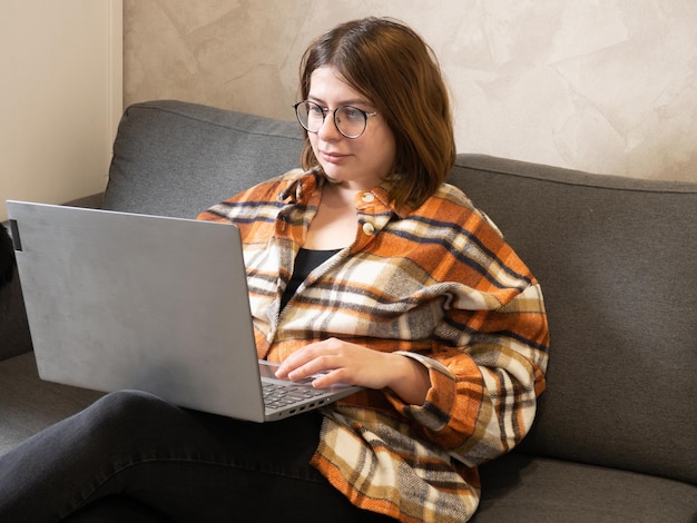 Jovem de óculos trabalhando em um laptop sentado em casa na cama.