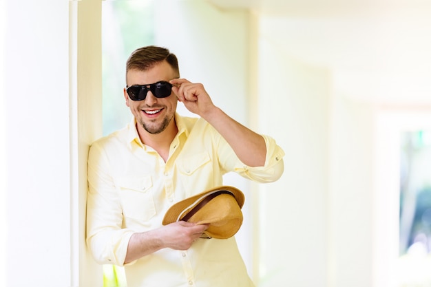 Jovem de óculos usando chapéu amarelo e roupas casuais se divertindo em dia de sol. Fundo tropical