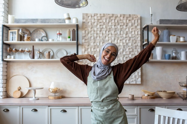 Jovem de origem oriental dona de casa em hijab em avental cozinha e limpa na cozinha ela dança