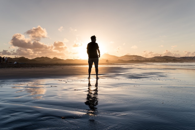 Jovem de pé em uma praia na maré alta ao pôr do sol.