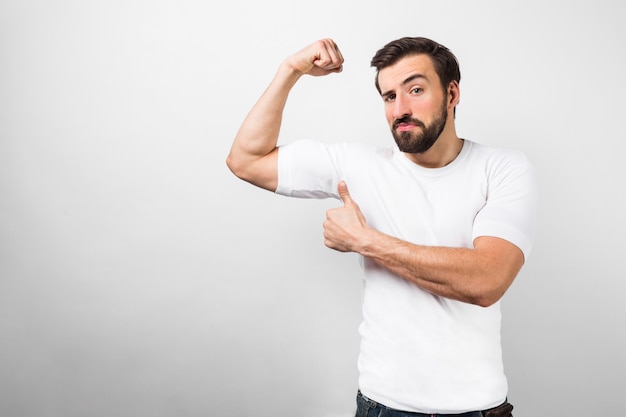 Jovem de pé isolado no fundo branco na camisa branca e mostrando um grande polegar na frente de seus músculos do braço. É porque o corpo dele está em boa forma e o cara sabe disso. fundo