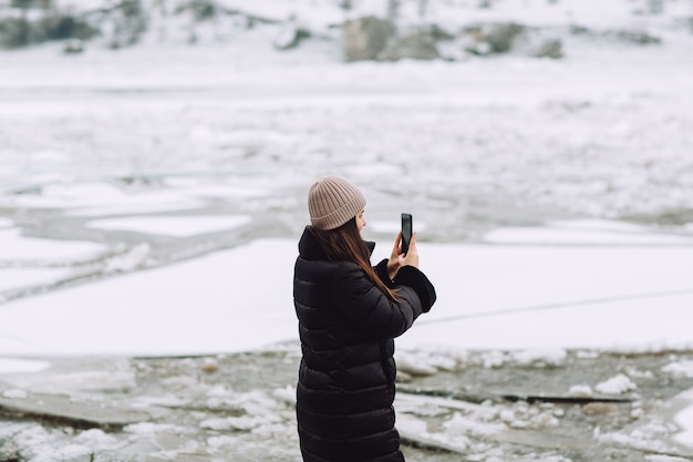 Jovem de pé na margem do rio coberta de blocos de gelo e tirando foto na paisagem nórdica.