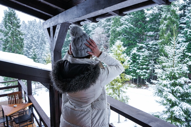 Jovem de pé no terraço em uma jaqueta branca e chapéu com vista para a floresta de inverno