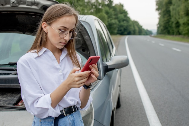 Jovem de pé perto de um carro quebrado com capô estourado, tendo problemas com seu veículo. Aguardando ajuda de reboque ou suporte técnico. Uma mulher liga para o centro de serviço.