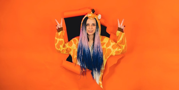 Jovem de pijama de girafa posando no buraco de fundo laranja Muito feminina em kigurumi olhando para a câmera e mostrando o gesto de paz