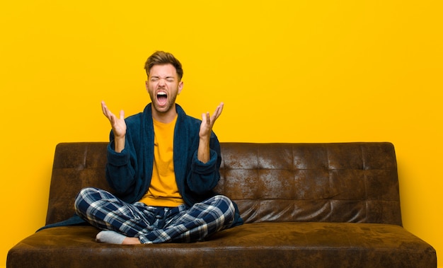 Jovem de pijama gritando furiosamente, sentindo-se estressado e irritado com as mãos no ar dizendo por que eu. sentado em um sofá