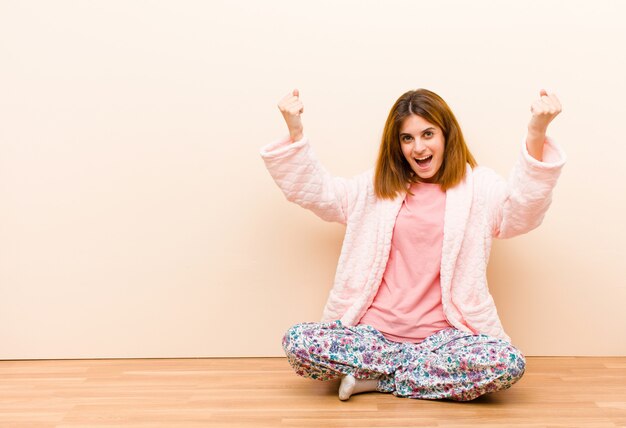 Jovem de pijama, sentada em casa, comemorando um sucesso inacreditável como um vencedor, parecendo animada e feliz dizendo, pegue isso!