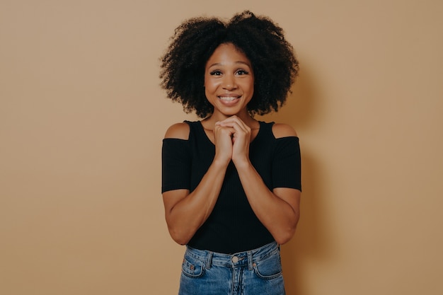 Jovem de raça mista de mãos dadas em gesto de oração, sorrindo com os olhos abertos isolados sobre um fundo bege escuro com espaço de cópia. mulher africana posando no estúdio, esperança e conceito de emoções positivas