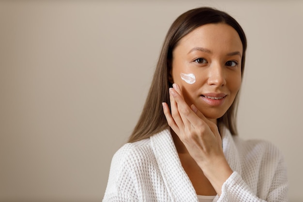 Foto jovem de roupão aplica loção ou hidratante no rosto cuidado facial profissional após a lavagem retrato de menina morena em fundo isolado