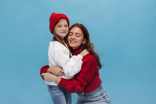 Jovem de suéter branco de chapéu vermelho e jeans abraça senhora sorridente em roupas brilhantes com olhos fechados em fundo isolado