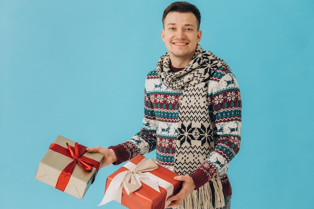 Jovem de suéter de Natal e cachecol segurando muitas caixas de presente com laço de fita de presente isolado em fundo azul Feliz ano novo conceito de celebração
