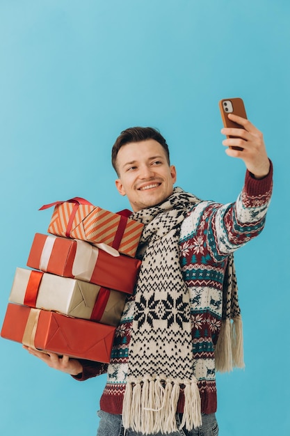 Jovem de suéter de Natal e cachecol segurando muitas caixas de presente e tirando selfie isolado em fundo azul Conceito de comemoração de feliz ano novo