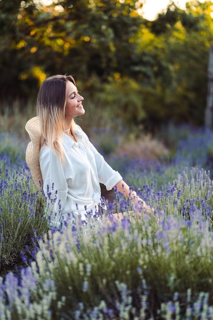 Jovem de terno de algodão branco e chapéu atrás das costas senta-se no campo de lavanda à noite