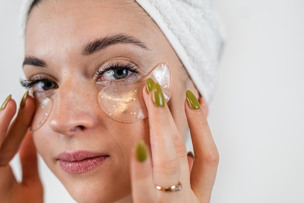 Jovem de toalha aplica manchas de beleza de hidrogel de colágeno sob os olhos isolados em branco