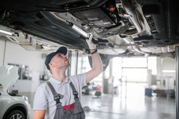 Jovem de uniforme fica debaixo do carro e olha para cima. Ele segura a luz na mão. Cara é sério e concentrado. Ele trabalha.