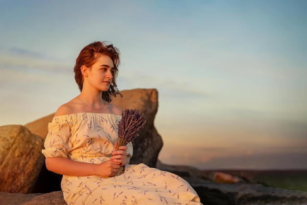 Jovem de vestido com buquê de lavanda nas mãos em uma colina no pôr do sol de verão Espaço para texto