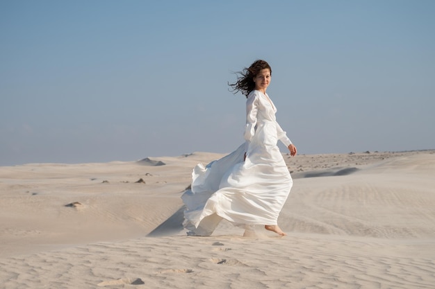 Jovem de vestido de noiva branco andando nas dunas de areia no deserto