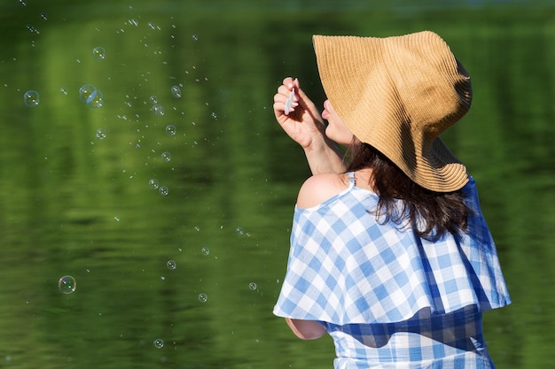 Jovem de vestido e chapéu de palha soprando bolhas no fundo do rio Vista traseira