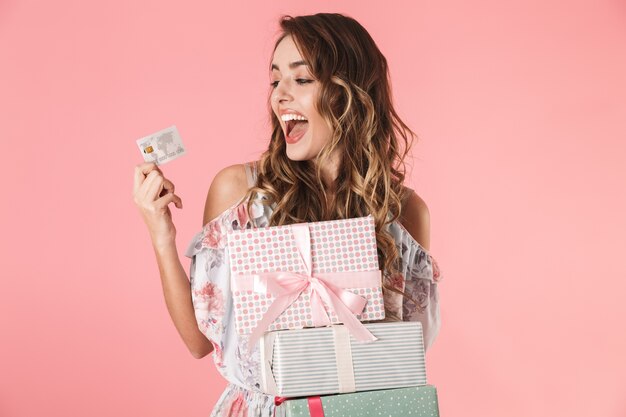 Jovem de vestido segurando um cartão de crédito e caixas com a compra, isoladas em rosa