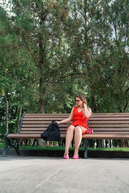 Jovem de vestido vermelho brilhante senta-se no banco no parque de verão Descanse no parque Quadro vertical