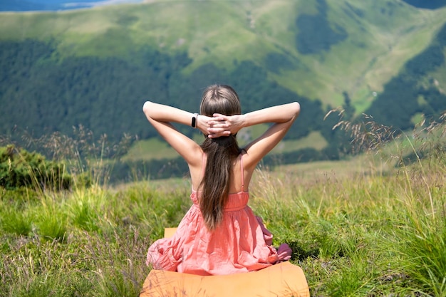 Jovem de vestido vermelho descansando no campo gramado verde em um dia ensolarado nas montanhas de verão apreciando a vista da natureza