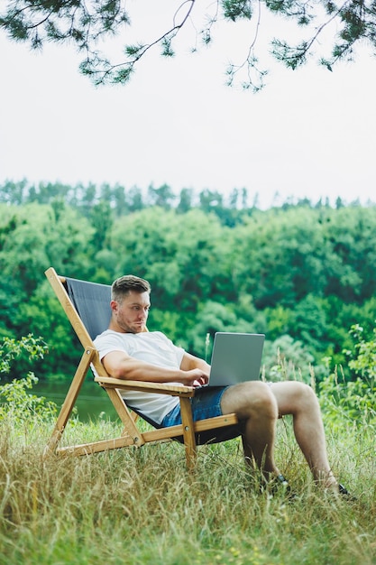 Jovem deitado em uma cadeira na natureza e trabalhando com um laptop descansando sozinho olhando pensativamente para cima Um homem em uma viagem no campo o conceito de estilo de vida das pessoas