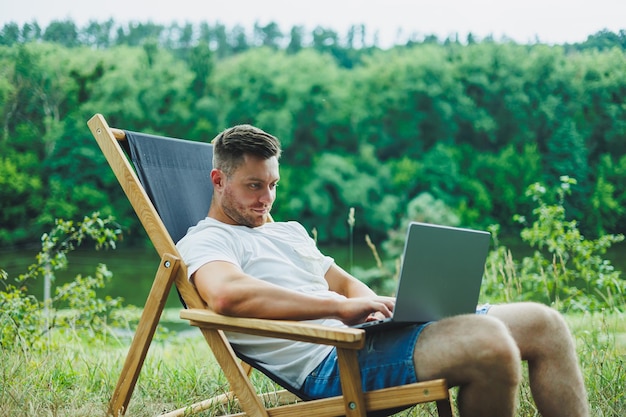 Jovem deitado em uma cadeira na natureza e trabalhando com um laptop descansando sozinho olhando pensativamente para cima Um homem em uma viagem no campo o conceito de estilo de vida das pessoas