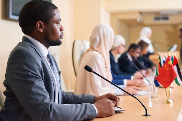 Jovem delegado afro-americano sério e participante de fórum político ouvindo relato de colega estrangeiro