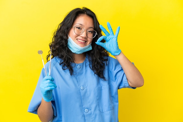 Jovem dentista asiático segurando ferramentas sobre fundo isolado, mostrando sinal de ok com os dedos