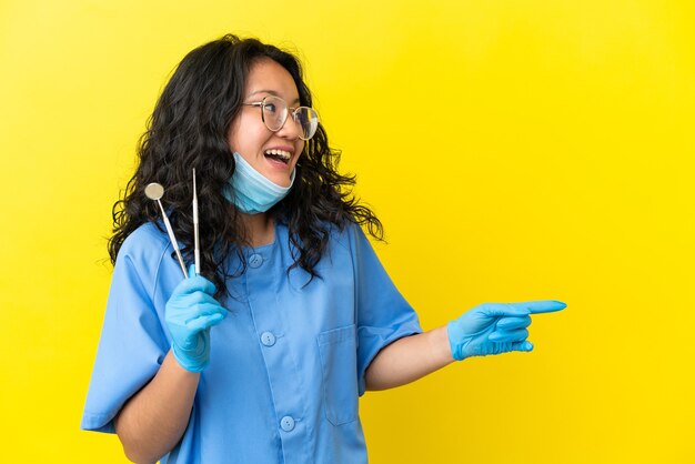 Jovem dentista asiático segurando ferramentas sobre um fundo isolado, apontando o dedo para o lado e apresentando um produto