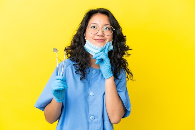 Jovem dentista asiático segurando ferramentas sobre um fundo isolado, pensando em uma ideia enquanto olha para cima