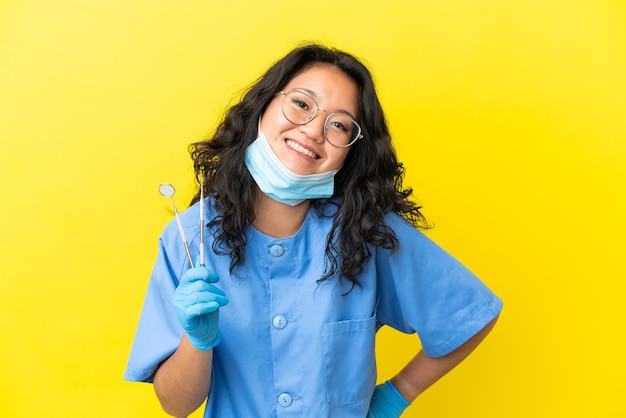 Jovem dentista asiático segurando ferramentas sobre um fundo isolado, posando com os braços na cintura e sorrindo