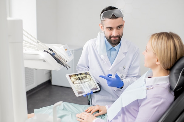 Jovem dentista bem sucedido em uniforme apontando para a tela sensível ao toque com raio-x de dentes enquanto mostra ao paciente o que deve ser feito com ela