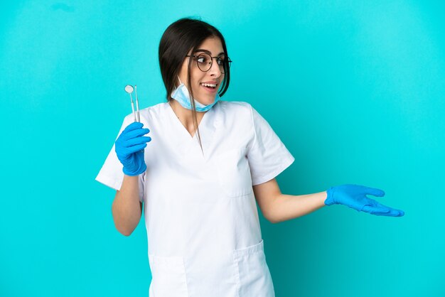 Jovem dentista caucasiana segurando ferramentas isoladas em um fundo azul com expressão de surpresa enquanto olha para o lado