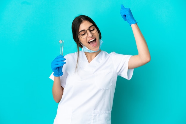 Jovem dentista caucasiana segurando ferramentas isoladas em um fundo azul, comemorando uma vitória