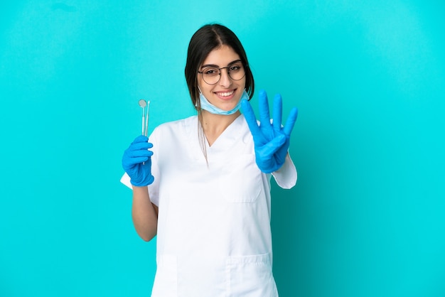 Jovem dentista caucasiana segurando ferramentas isoladas em um fundo azul feliz e contando quatro com os dedos