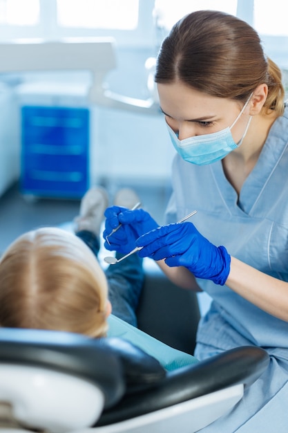 Jovem dentista simpática segurando uma sonda dentária e um espelho bucal e prestes a fazer um check-up na cavidade oral de seu pequeno paciente