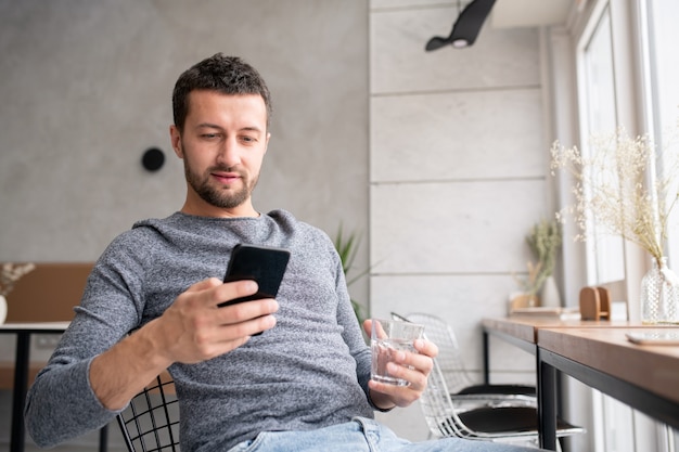 Jovem descansado relaxando em um café aconchegante, tomando um copo d'água e rolando ou enviando mensagens no smartphone