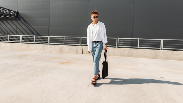 Jovem descolado americano com roupas elegantes em sandálias vermelhas com uma bolsa de tecido em óculos de sol está na rua, perto do prédio cinza. Cara na moda ao ar livre. Nova coleção de moda masculina de verão.