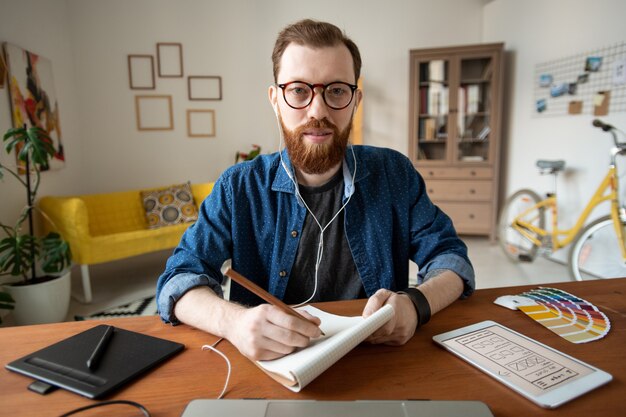 Foto jovem desenvolvedor de software criativo barbudo com fones de ouvido fazendo anotações ou esboços na página do bloco de notas na frente do laptop em casa