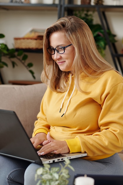 Jovem desenvolvedora de software feminina muito sorridente na glasse, trabalhando em um laptop em casa