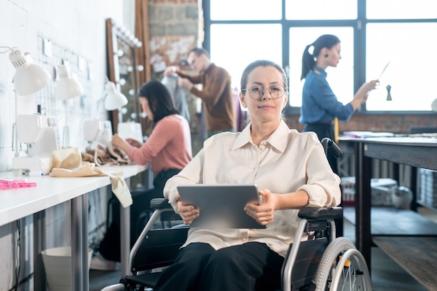 Jovem designer criativo usando um tablet enquanto está sentado em uma cadeira de rodas com colegas trabalhando em uma nova coleção de moda em uma grande oficina