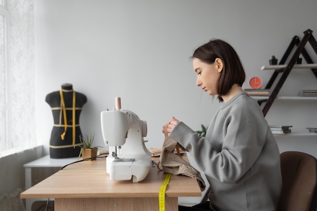 Jovem designer de moda caucasiana está sentado em casa em seu escritório à mesa Máquina de costura Meça o pedaço de tecido necessário com um medidor flexível Conceito de pequena empresa