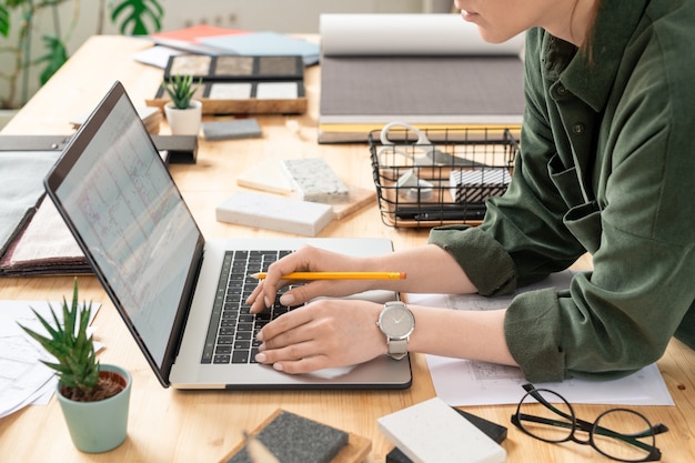 Jovem designer feminina criativa, curvada sobre a mesa em frente ao laptop enquanto trabalhava com o desenho eletrônico de uma casa ou apartamento perto do local de trabalho