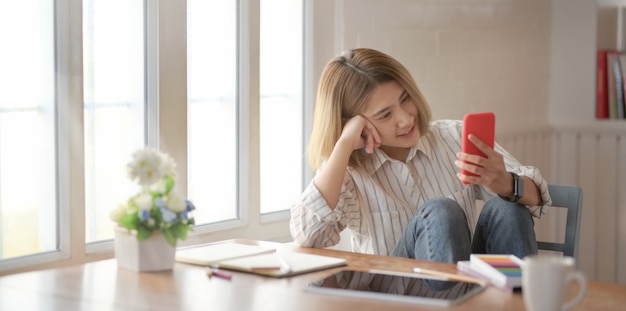 Jovem designer feminino bonito olhando para smartphone e sentado na cadeira