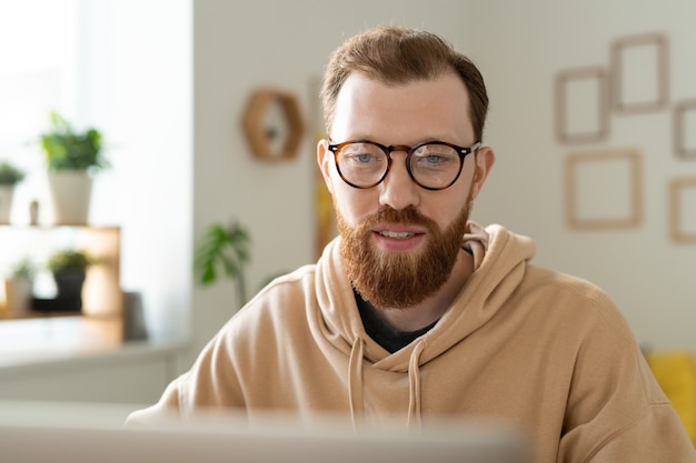 Jovem designer freelance barbudo usando óculos, olhando para a tela do laptop enquanto rola as informações on-line durante o trabalho remoto