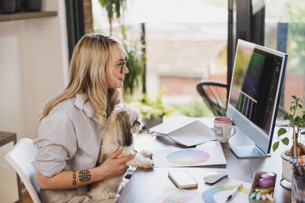 Jovem designer freelancer atraente trabalhando no computador em sua casa e tendo seu cachorro de estimação no colo para lhe fazer companhia.