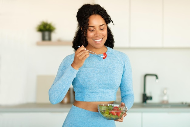 Jovem desportista afro-americana comendo salada de legumes fresca na cozinha desfrutando de comida cheia de vitaminas copie o espaço
