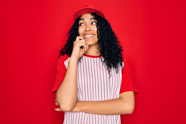 Jovem desportista afro-americana encaracolada usando boné de beisebol e camiseta listrada com mão no queixo pensando em pergunta expressão pensativa Sorrindo com rosto pensativo Conceito de dúvida